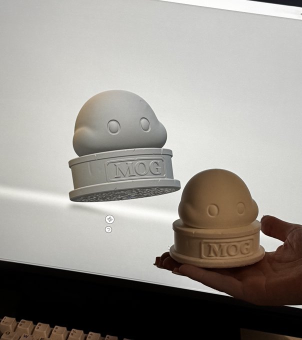 A 3D-printed "MOG" mascot figurine is held up next to its digital design displayed on a computer screen, showcasing the tangible product created by the couple beyond just online guides and threads.
