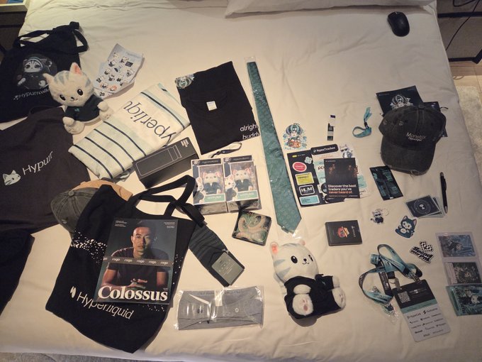 An assortment of merchandise and items is laid out on a white bed, including a 'Colossus' magazine, 'Hyperliquid' branded clothing, plush toys, stickers, and a laptop.