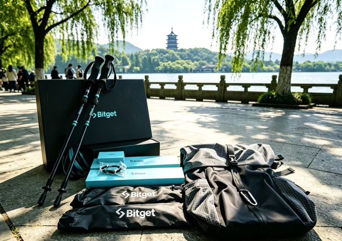 Outdoor display of Bitget-branded merchandise including a black box, trekking poles, a jewelry box, a gray duffel bag, and a black jacket laid out on stone paving near a lake with a pagoda in the background.