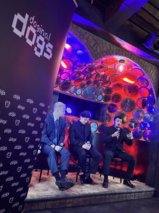 Three men in suits sit on a stage at a 'dosi/dogs' event, with one speaking into a microphone in front of a colorful circular decorative backdrop.