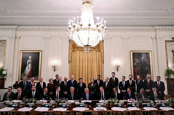 A group photo of government officials and politicians posing together at a long table in an ornate room with chandeliers and historical portraits, likely a White House cabinet meeting.