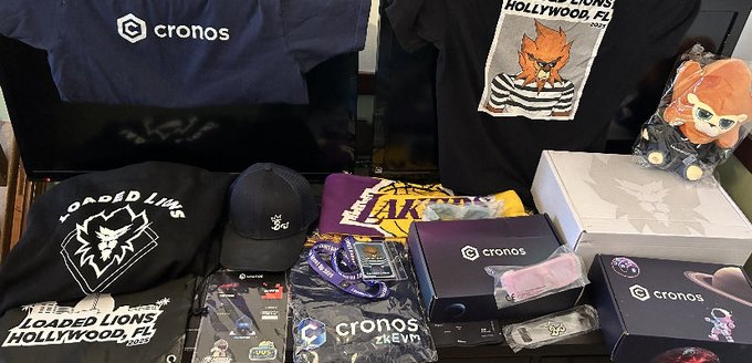 A Crypto.com branded merchandise haul featuring "cronos" apparel, a "Loaded Lions Hollywood" t-shirt, accessories, and collectible figurines sits displayed, seemingly shared as a comparison to the user's frustration over never receiving their upgraded Crypto.com card.