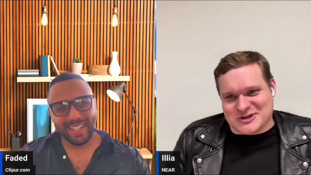 The image shows two men sitting next to each other, with one man wearing a leather jacket and glasses while the other has a black shirt and sunglasses. They are both smiling at the camera, appearing to be engaged in a conversation or discussion. The background of the image is white, which contrasts with the brown wooden wall behind them. There are also several books visible on the wall, suggesting that they might be discussing a topic related to literature or research.
