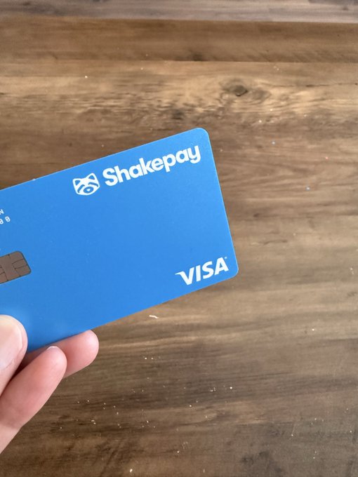A hand holds up a blue Shakepay Visa debit card against a wooden surface background.