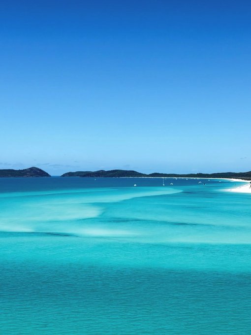 A serene photograph of a tropical beach landscape, showcasing crystal-clear turquoise waters with visible white sandbars, a distant shoreline with hills, and a clear blue sky.