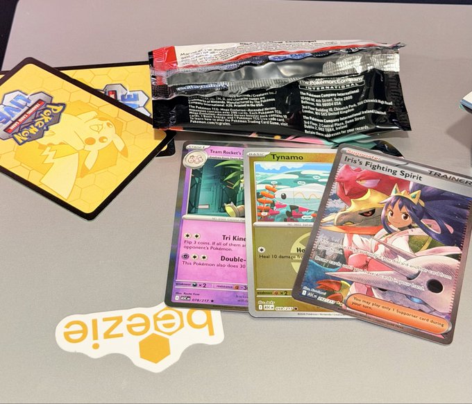 A collection of Pokémon TCG items including a sealed booster pack, cards featuring Team Rocket's Tri King, Tynamo, and 'Iris's Fighting Spirit,' along with a beehive logo sticker and branded card sleeves.