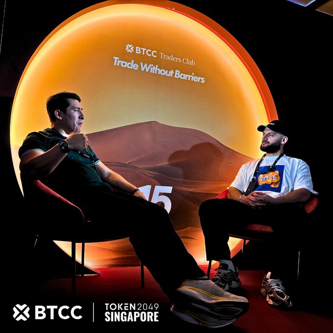 Two men seated in a discussion panel on a stage with an orange sun-like backdrop, branded 'BTCC Traders Club Trade Without Barriers' at the TOKEN2049 SINGAPORE event.