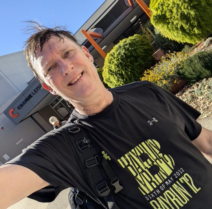 A young man in a black t-shirt featuring the "Bay to Bayrun 12" running event graphic takes a selfie outdoors near a building labeled "ORANGE LEES."