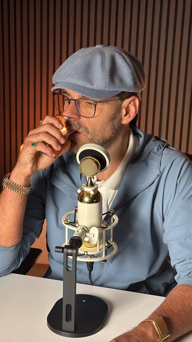 A man wearing a gray newsboy cap and light blue jacket drinks from a glass while sitting in front of a studio microphone, seemingly mid-podcast recording. The image ties to the post by playfully suggesting the host is taking a sip—drinking something other than the stock—before declaring he's not buying $LAPTOP that day.
