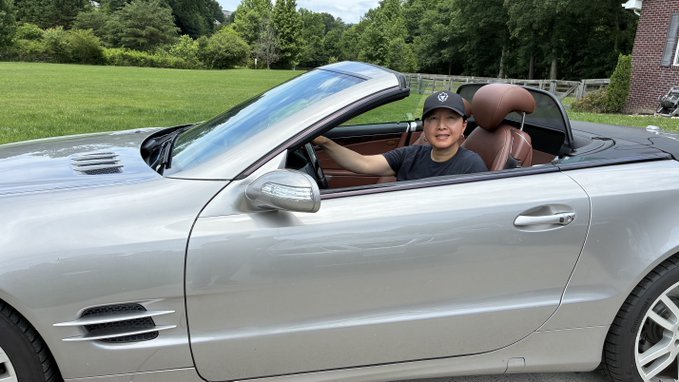 The image features a man sitting inside a silver convertible car, wearing a black hat for sun protection while driving down the street. The car has an open top design, allowing the driver to enjoy the surrounding environment. The caption of the image includes the hashtag #LOADEDLIONS and mentions that it is an NFT from the Flex Your Loaded Merch company.