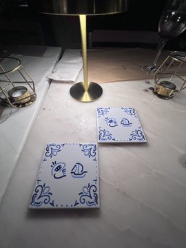 The image features a dining table with two small white ceramic tiles featuring blue designs of boats sitting on top of it. The table also has a gold lampshade and a wine glass placed nearby. In the background, there are other tables and chairs, suggesting that this is an event or gathering taking place in a restaurant setting.