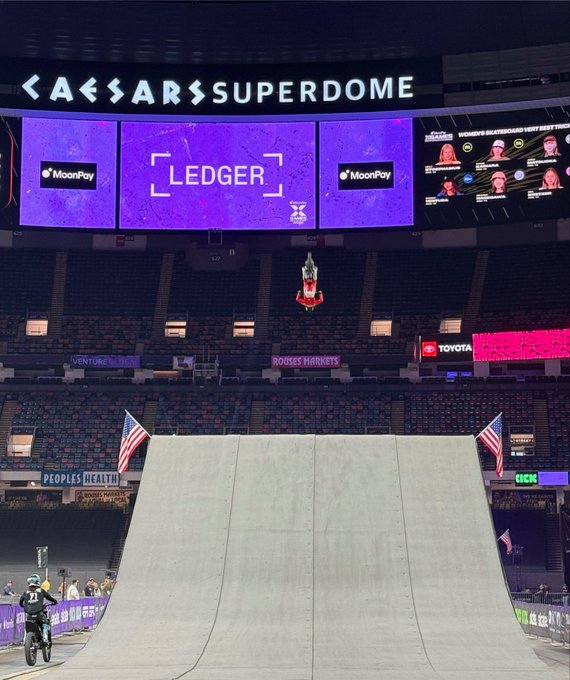 A motocross rider is mid-air executing an inverted backflip over a massive ramp inside the Caesars Superdome, with the venue's jumbotron displaying "LEDGER," "MoonPay," and "Toyota" sponsor logos alongside the text "MONDAY NIGHT RAW" — capturing the focus and precision that matches the caption "so locked in."