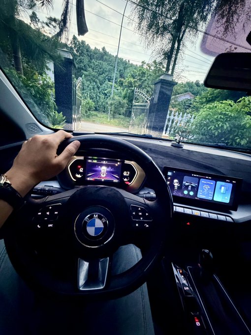 Interior view from the driver's seat of a BMW, showing the steering wheel with the BMW logo, digital dashboard, and infotainment screen, with a gated entrance visible through the windshield.