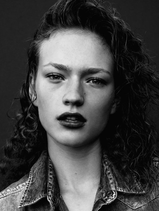 A black-and-white studio portrait shows a woman with curly hair, dark lipstick, and a denim jacket facing the camera.