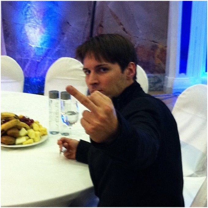 A young man with brown hair, wearing a dark sweater, points his finger at the camera while sitting at a table with a plate of snacks and two bottles of water in a room lit with blue lighting. The image relates to the post as it likely depicts Pavel Durov, the founder of Telegram, who is the subject of Russia's international arrest warrant mentioned in the accompanying text.