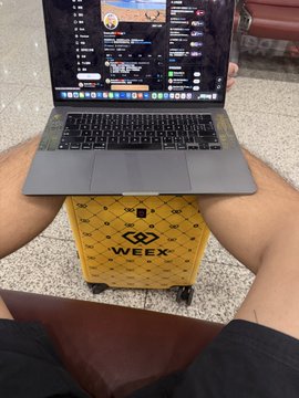 The image shows a man sitting down with his legs crossed, holding an open laptop computer that has the word "WEEX" written on its side. The laptop is placed on top of a yellow suitcase or luggage bag, which appears to be a Weex luggage container. The scene takes place in what seems to be an airport setting, as indicated by the presence of a chair and other airport-related objects such as a backpack and a handbag.