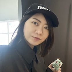 The image features an Asian woman wearing a black baseball cap with white lettering that reads "IN". She is holding up a small toy Pokemon figure against a dark background, possibly posing for a photo or video. The caption below the image provides information about the person and their role in the organization, which appears to be related to Pokemon.