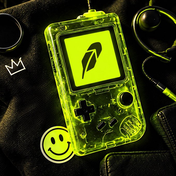A translucent neon yellow Game Boy Color featuring a feather quill icon on the screen, styled alongside headphones, a smiley face sticker, and a crown logo on a dark background.