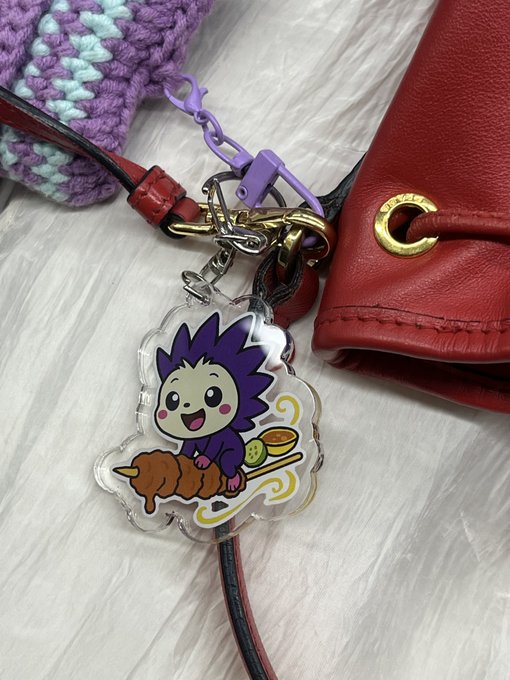 An acrylic charm keychain featuring a cute purple-haired cartoon character sitting on what appears to be grilled meat with a side dish, attached to a red leather bag.