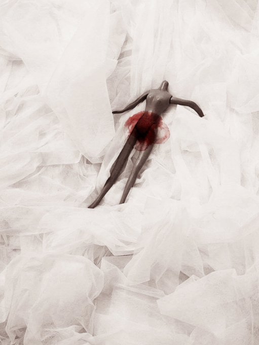Dark mannequin partially wrapped in white tulle/fabric with a prominent red blood stain at the midsection, set against a draped white fabric background.