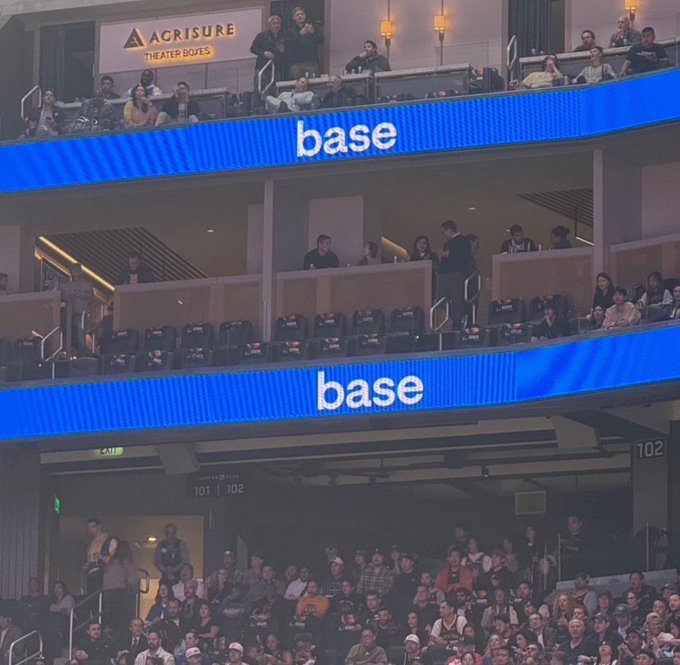 The image shows a large crowd of people sitting in chairs inside an arena or stadium. They are watching something happening on stage, possibly during a sports event. The audience appears engaged and focused on the action taking place. The text "Slowly then all at once" is visible in the image, which could be related to the post about the @base.
