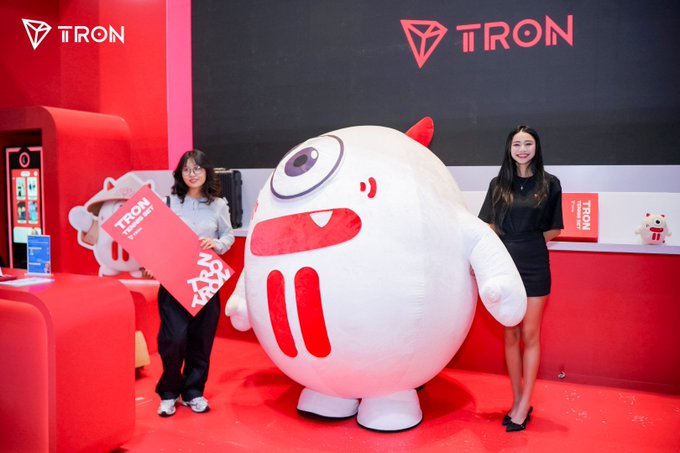 Two women pose with a large white mascot featuring a single eye at the TRON (TRX) branded booth, with one holding a red promotional item, promoting TRON's presence at the Conviction event in Ho Chi Minh City.