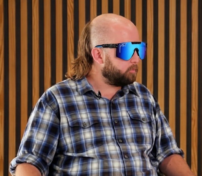 A man with a shaved head, beard, and long hair at the back, wearing blue reflective Pit Viper sunglasses and a blue plaid shirt.