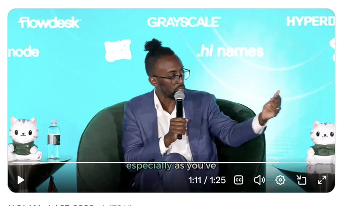 A man in a blue blazer speaks into a microphone at a conference panel, with sponsor logos including "Flowdesk," "Grayscale," "Hyperliquid," and ".hi names" displayed on the backdrop behind him, connecting to the post's reference to Hyperliquid. The video timestamp shows 1:11 of 1:25, with on-screen text reading "especially as you've."