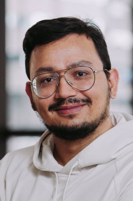 The image features a man wearing glasses, a white hoodie, and black hair standing against a gray background. The picture's caption discusses Polygon's total stablecoin supply and volume as well as stablecoins' value in transactions. 

In the image, the man is staring directly at the camera with a neutral expression on his face, which adds an interesting dynamic to the scene.