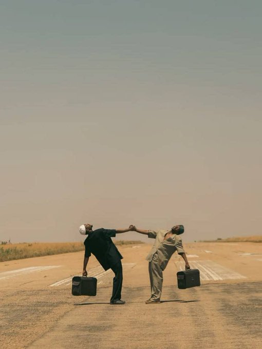 A cinematic photograph of two men in suits standing on an empty desert road, leaning backward with outstretched arms as if being pulled apart, each holding a briefcase, against a hazy pale sky.
