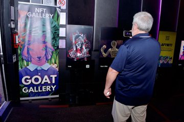 A man in a blue polo shirt viewing digital artwork at the 'GOAT GALLERY' NFT exhibition, standing in front of screens displaying animated NFT pieces.