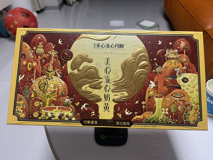 An ornate gold and red mooncake gift box from 美心 (Meixin) featuring a phoenix/bird design and Chinese characters, displayed on a CoinUp.to scale.