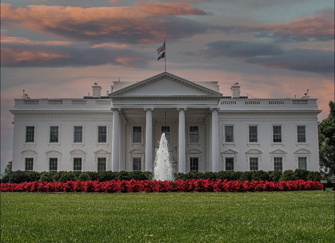 The White House at sunset with the American flag flying atop, surrounded by red flowers and a fountain in the front lawn.