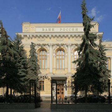 The image shows the Bank of Russia headquarters, a grand neoclassical building with "БАНК РОССИИ" (Bank of Russia) inscribed across its facade, flying the Russian flag. This connects to the announcement as the central bank is publishing its first framework for regulating organized crypto trading, setting capital requirements and custody rules for digital asset depositories.
