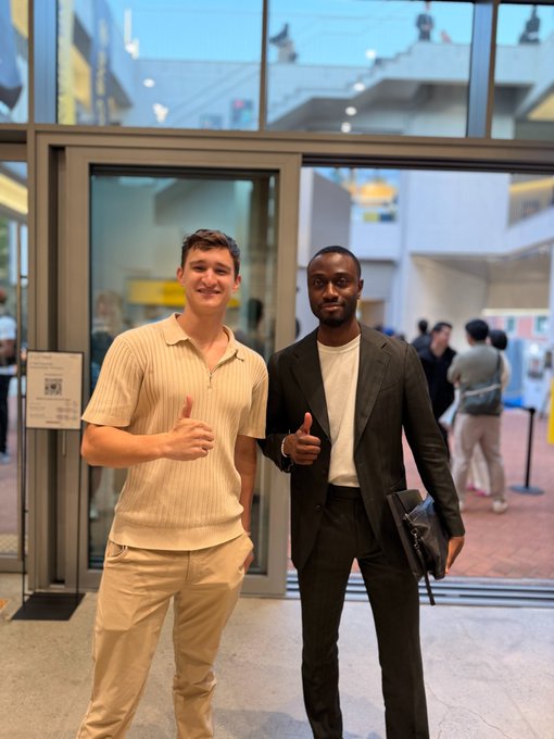Two men stand indoors near a glass entrance, both giving a thumbs-up; one wears a striped polo and the other a black blazer.