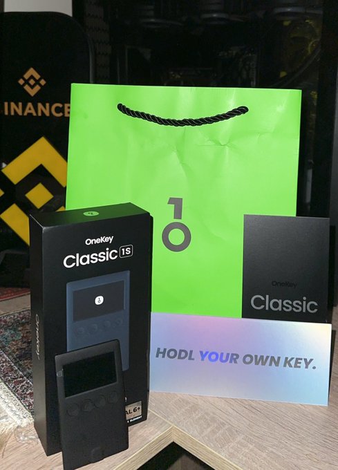 The image shows a OneKey Classic 1S hardware wallet in its black packaging alongside its user manual and a bright green shopping bag, with the tagline "HODL YOUR OWN KEY" visible. The setup corresponds perfectly with the post's acknowledgment of receiving the OneKey hardware wallet as a gift from the @OneKeyHQ team in 2024.