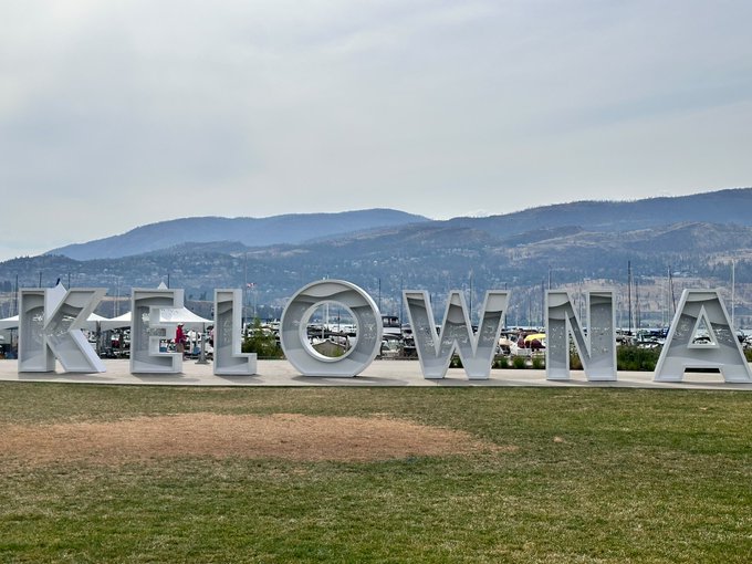 Large silver letters spelling 'KELOWNA' stand on a grassy area with mountains and a parking lot visible in the background.
