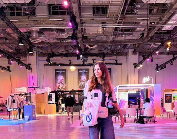 A woman stands in the center of a large, modern event hall with pink and purple lighting, holding a white tote bag, with various booths and screens in the background.