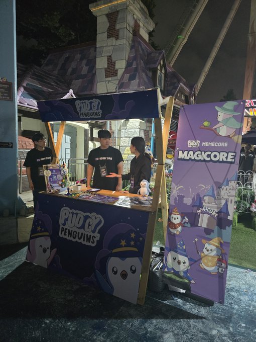 A nighttime view of a merchandise booth at an event called 'Magicore,' featuring 'Pudgy Penguins' branding with purple banners showing cartoon penguin mascots in wizard attire.