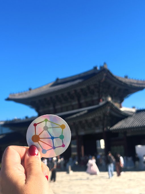 A hand with red nail polish holding a holographic sticker with the Sui (SUI) network logo in front of a traditional East Asian temple under a clear blue sky.