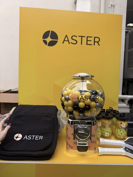 A promotional display features the brand 'ASTER' on a yellow backdrop, with a gumball machine filled with yellow and black balls, branded bags, and drinkware.