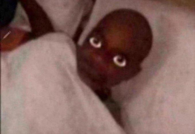 A close-up, slightly blurry image of a person with dark skin wrapped in white fabric or bedding, looking toward the camera with wide eyes.