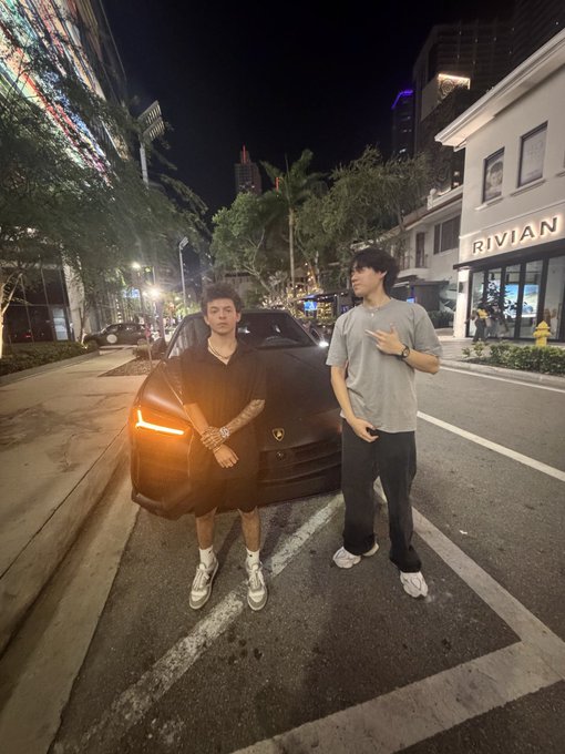 Two young men pose confidently at night on a city street beside a black Ferrari with illuminated headlights, while the illuminated "RIVIAN" storefront glows in the background—capturing a "I'll be back" vibe of return and reunion with luxury and style.