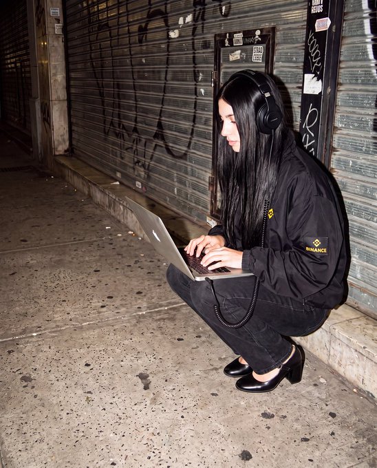 A young woman with long black hair wearing a Binance branded jacket squats on a sidewalk at night while typing on a silver laptop, wearing large black headphones in front of closed graffiti-covered metal shutters.