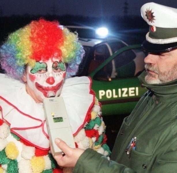 A clown with a rainbow wig and makeup blows into a breathalyzer held by a uniformed German Polizei officer, with a police car visible in the background.