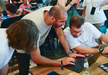 Two men lean intently over a laptop at the Web3 Summit 2026, collaborating closely as they examine something on the screen, with other attendees visible in the background of the busy event space. The image captures the hands-on, collaborative spirit of the Builder's Impact Award and Open Knowledge Award ceremony, where PBA recognized projects advancing Web3's real-world potential and accessibility.