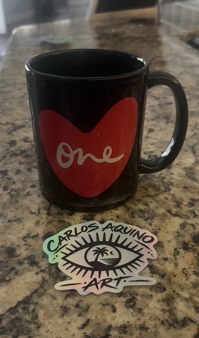 A black ceramic mug featuring a red heart with the word 'One' sits on a granite countertop next to a sticker labeled 'Carlos Aquino Art'.