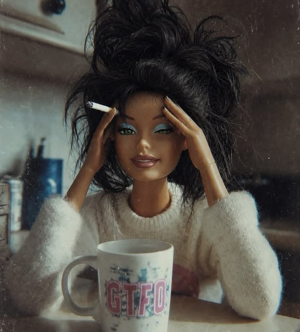 A Bratz-style doll with messy black hair, blue eyeshadow, and a fuzzy white sweater holds a cigarette while sitting at a table with a mug that reads 'GTFO'.