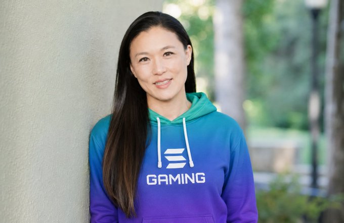 A smiling woman with long dark hair leans against a wall, wearing a blue and purple hoodie featuring the "E" logo and the word "GAMING"—the account holder presenting herself as the person behind the brand.