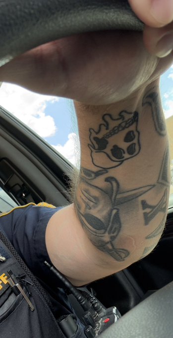 A person's forearm displays a Bored Ape Yacht Club (BAYC) tattoo featuring the iconic cartoon ape with a crown and bandana, visible while they sit in a vehicle wearing a police-style uniform. The tattoo directly reflects the post's reference to "BAYC," showcasing the wearer's personal connection to the NFT collection.